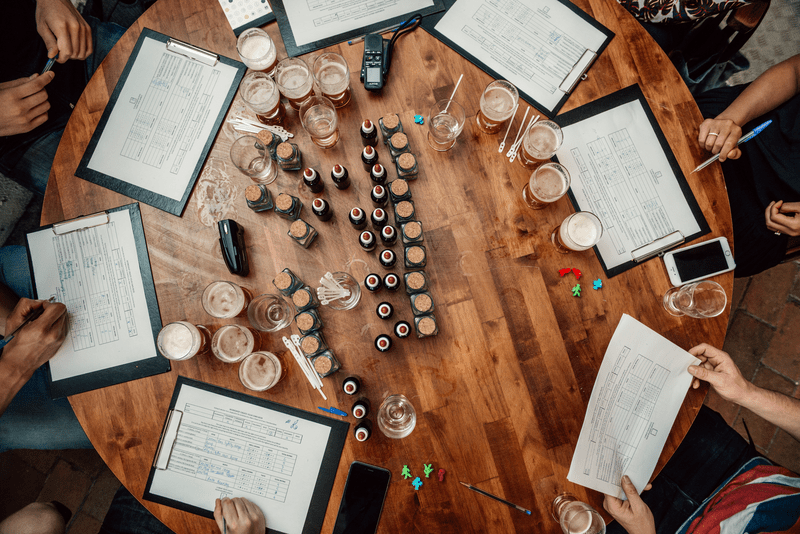 Vista aérea de una mesa redonda de madera con portapapeles, vasos pequeños y botellas. Las personas están sentadas alrededor, sosteniendo bolígrafos, lo que sugiere una sesión de degustación.