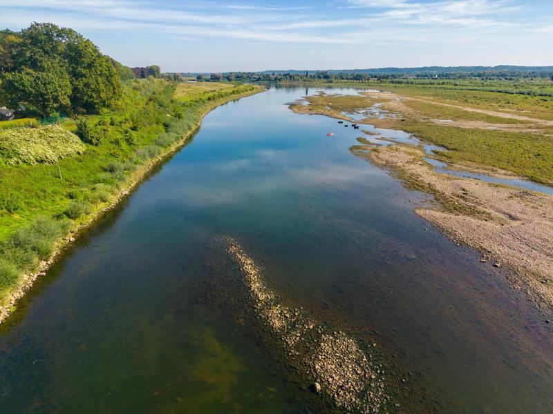 Een sereen rivieroeverlandschap met een kronkelende rivier, omringd door weelderig groen en kiezelige oevers onder een helderblauwe lucht. Rustig en uitgestrekt.