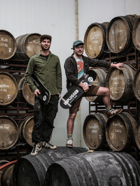 Alex Lippens & Koen Van Lancker Twee mannen met skateboards poseren tussen gestapelde houten vaten in een brouwerij- of distilleerderijmagazijn.
