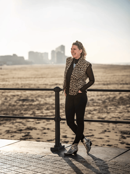 Shannah Zeebroek Woman leaning against a railing on the promenade, with sandy beach and a blurred city skyline in the background.