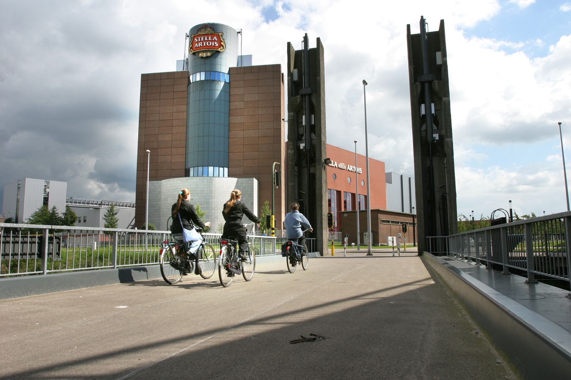 Drei Personen fahren mit dem Fahrrad über eine Brücke auf ein hohes, modernes Gebäude mit einem Stella-Artois-Schild zu, unter einem teilweise bewölkten Himmel. Eine urbane, dynamische Szenerie.
