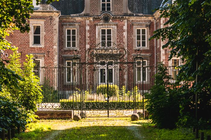 Een statig bakstenen herenhuis omgeven door weelderig groen, met zonnestralen die door de bomen vallen. De lucht is helder, wat een rustige en uitnodigende sfeer creëert.