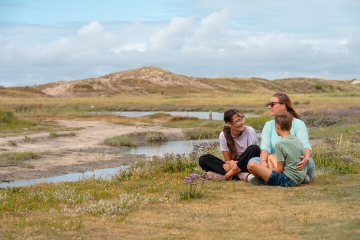 Parc paysager flamand de la région de Zwinstreek Une famille est assise sur des dunes herbeuses près d’un ruisseau sous un ciel nuageux, profitant d’un moment paisible. La scène transmet chaleur, détente et convivialité.