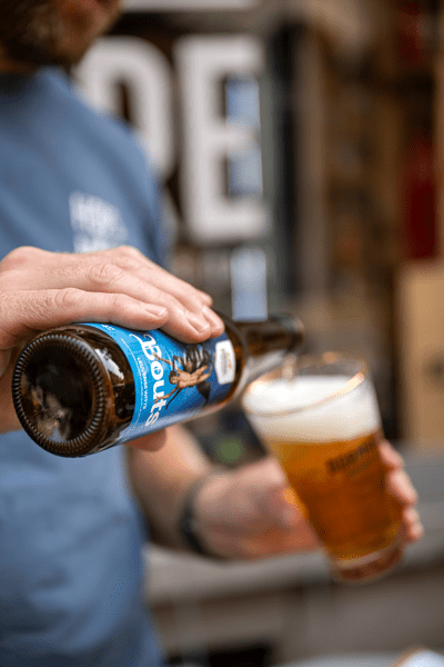 A person pours beer from a bottle with a blue label into a glass, creating frothy foam. The atmosphere is casual and relaxed.