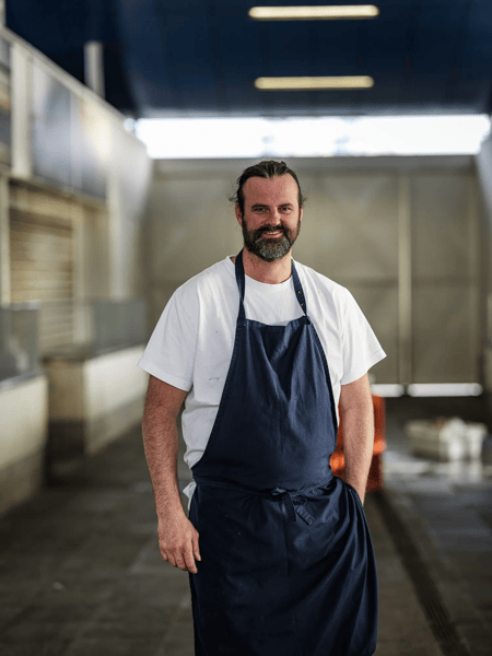 Willem Hiele Bearded man in a white T-shirt and dark apron stands in an industrial kitchen, looking at the camera.