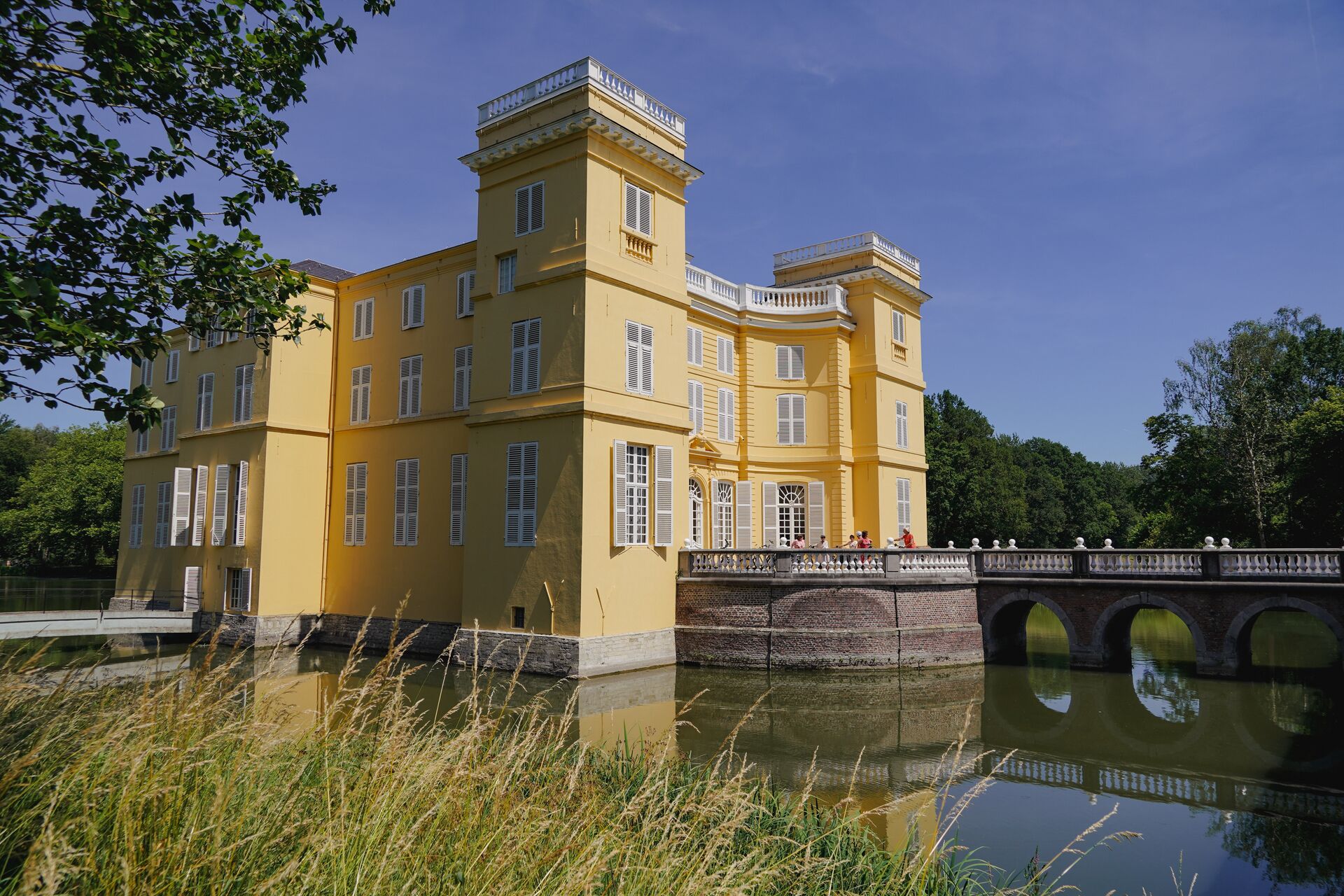 Kasteel d'Ursel con puente junto al agua Castillo amarillo junto al agua con puente de piedra y reflejo, rodeado de árboles bajo cielo azul.