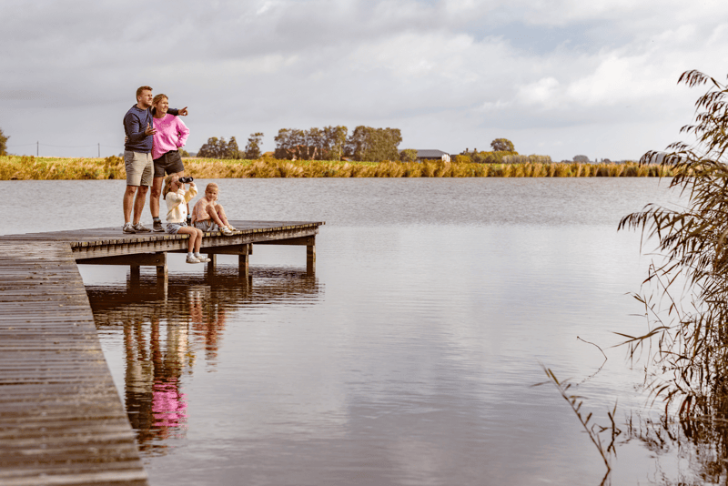 A family with a dog enjoys a calm lake view from a wooden dock under a cloudy sky. A child sits on the dock edge, creating a serene, peaceful scene.