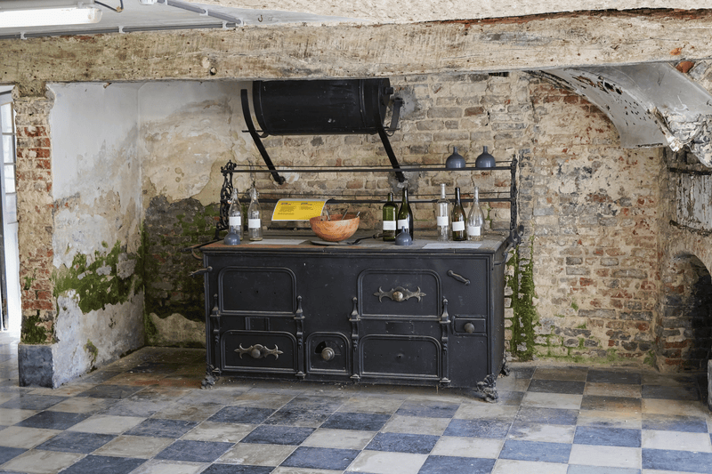 Rustieke keuken met een oude zwarte ijzeren stove tegen een verweerde bakstenen muur. Op de stove staan wijnflessen en een houten kom, wat een historische sfeer oproept.