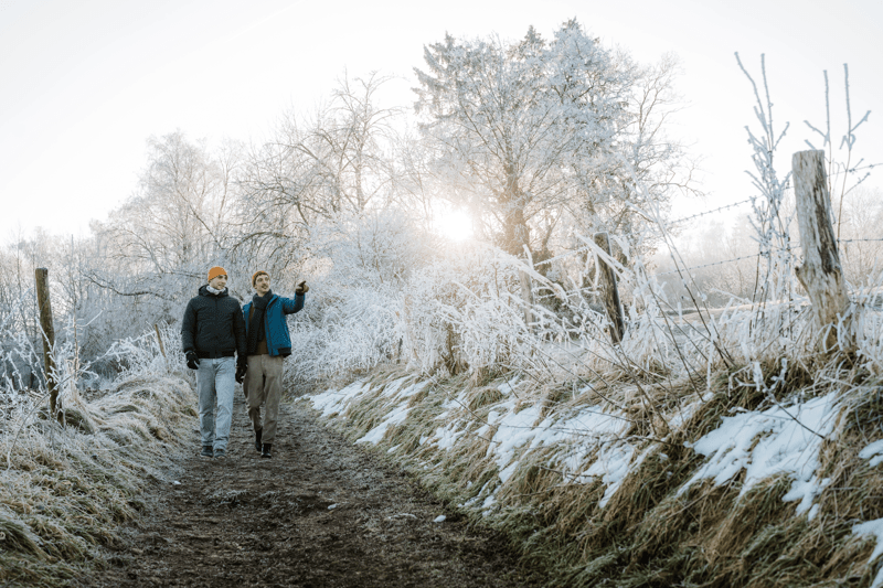 Dos personas caminan por un sendero helado bordeado de árboles y hierba cubiertos de nieve. El amanecer proyecta un cálido resplandor que transmite una tranquila mañana de invierno.