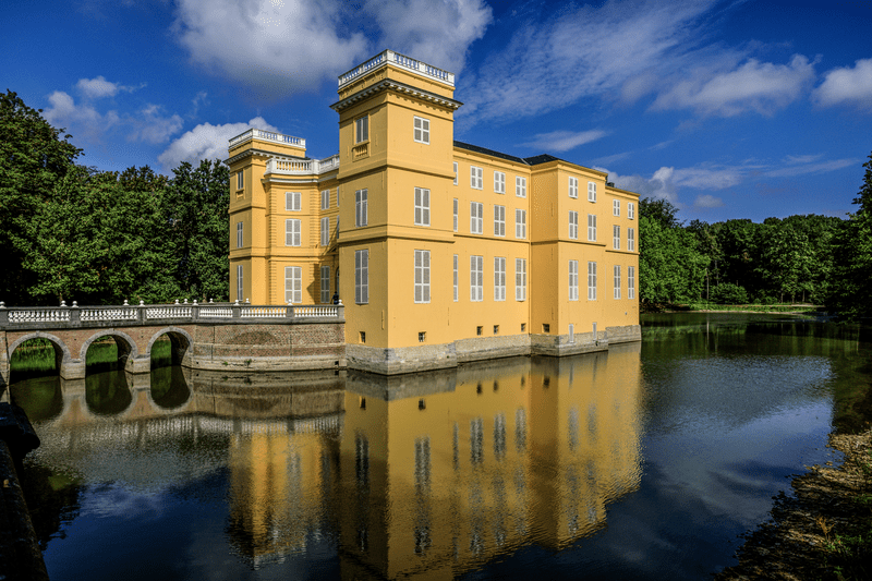 Kasteel d’Ursel Een geel kasteel met hoge torens en grote ramen weerspiegelt zich in een omliggende gracht. De lucht is blauw met enkele wolken en er zijn bomen in de buurt zichtbaar.