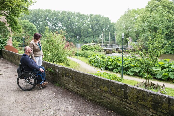 Een persoon in een rolstoel en een begeleider genieten van een rustig, schilderachtig uitzicht op een weelderig park met bomen en een rivier, wat rust en verbondenheid uitstraalt.