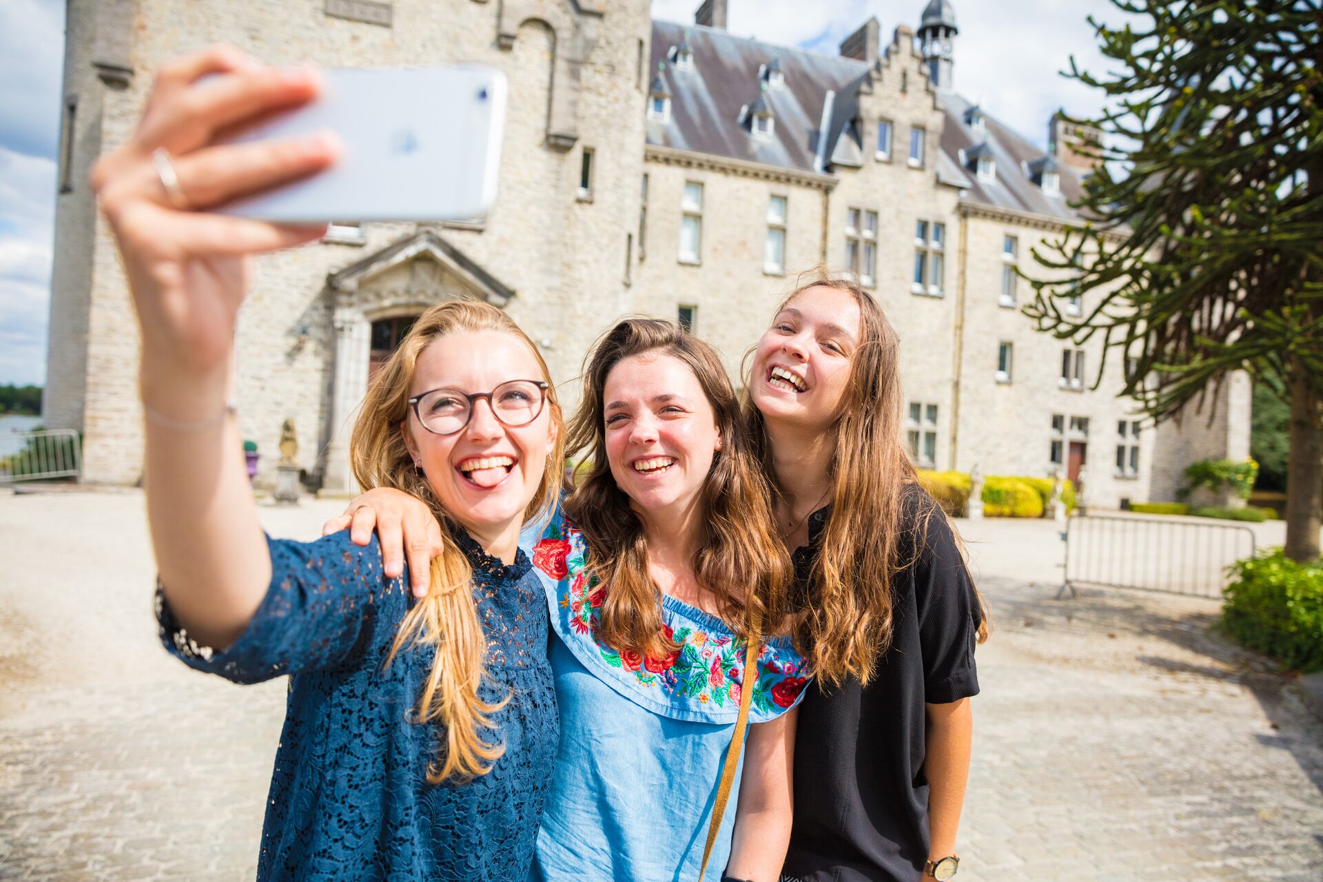 Tre amici si scattano un selfie con gioia davanti a un edificio storico in pietra con finestre ad arco. Sorridono felici sotto il sole.