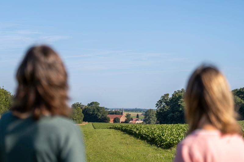 Twee personen met wazig haar kijken uit over een dorp in de verte, omringd door weelderige groene velden en bomen onder een helderblauwe lucht, wat een gevoel van rust oproept.