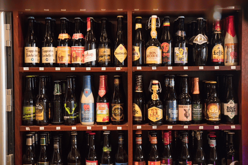Holzschrank mit Reihen belgischer Bierflaschen mit verschiedenen Etiketten wie Rochefort, Westmalle und Leffe.
