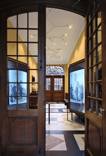 Museum Plantin Moretus Doorkijk door glazen houten deuren naar verlichte gang met vitrines en zwart-witte illustraties.