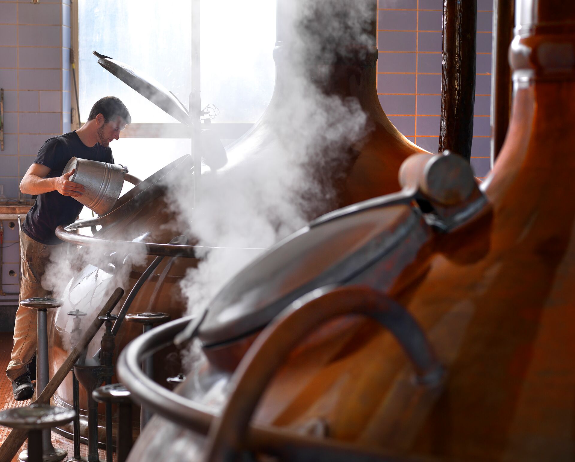 Brouwerij Het Anker, Mechelen Eine Person gießt sorgfältig Flüssigkeit in einen dampfenden Behälter in einer Brauerei. Das warme Licht und der Dampf schaffen eine konzentrierte und industrielle Atmosphäre.