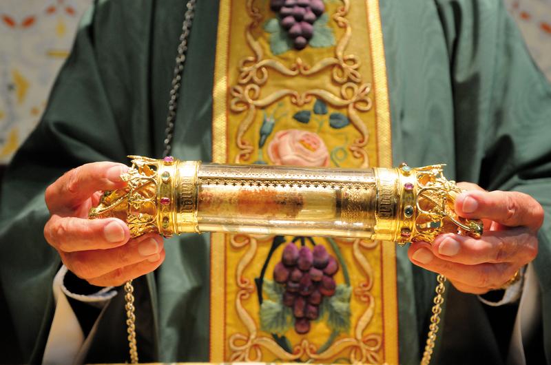 Een persoon in sierlijke groene gewaden met bloemrijke borduursels houdt een ingewikkeld glazen reliekschrijn met gouden rand, wat eerbied en traditie uitstraalt.