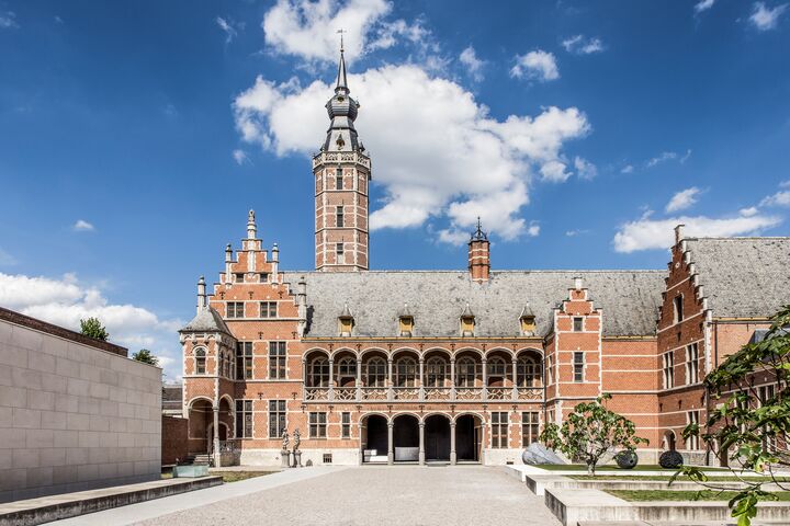 Museum Hof van Busleyden in Mechelen Historisch gebouw met rode bakstenen gevel en sierlijke toren onder een helderblauwe lucht met witte wolken. Een serene sfeer met een ruime binnenplaats.