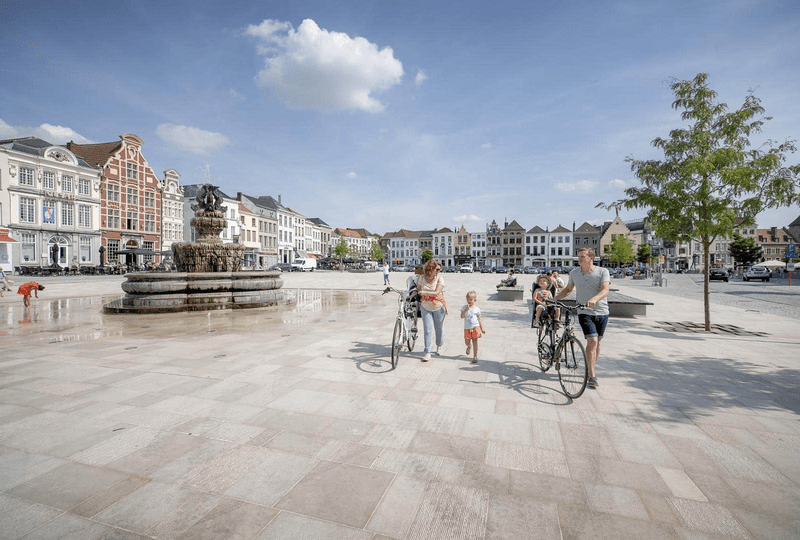 Une famille fait du vélo à travers une grande place ensoleillée avec une fontaine centrale. Des bâtiments historiques bordent l’arrière-plan, évoquant une ambiance animée et détendue.