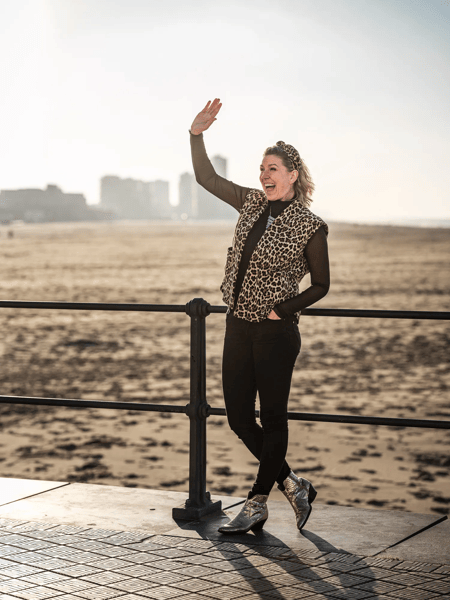 Shannah Zeebroek Woman in a leopard-print jacket waves by a railing on the boulevard, with beach and a hazy skyline in the background.
