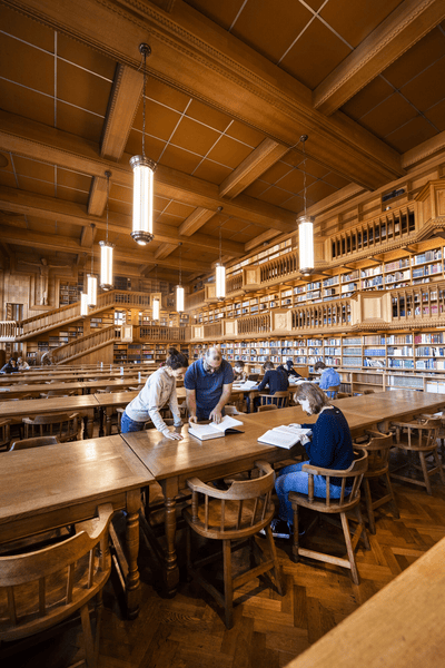 Grote Leeszaal in de Universiteitsbibliotheek, Leuven Grote houten leeszaal van een bibliotheek met lange tafels, boekenrekken en mensen die studeren onder hanglampen.