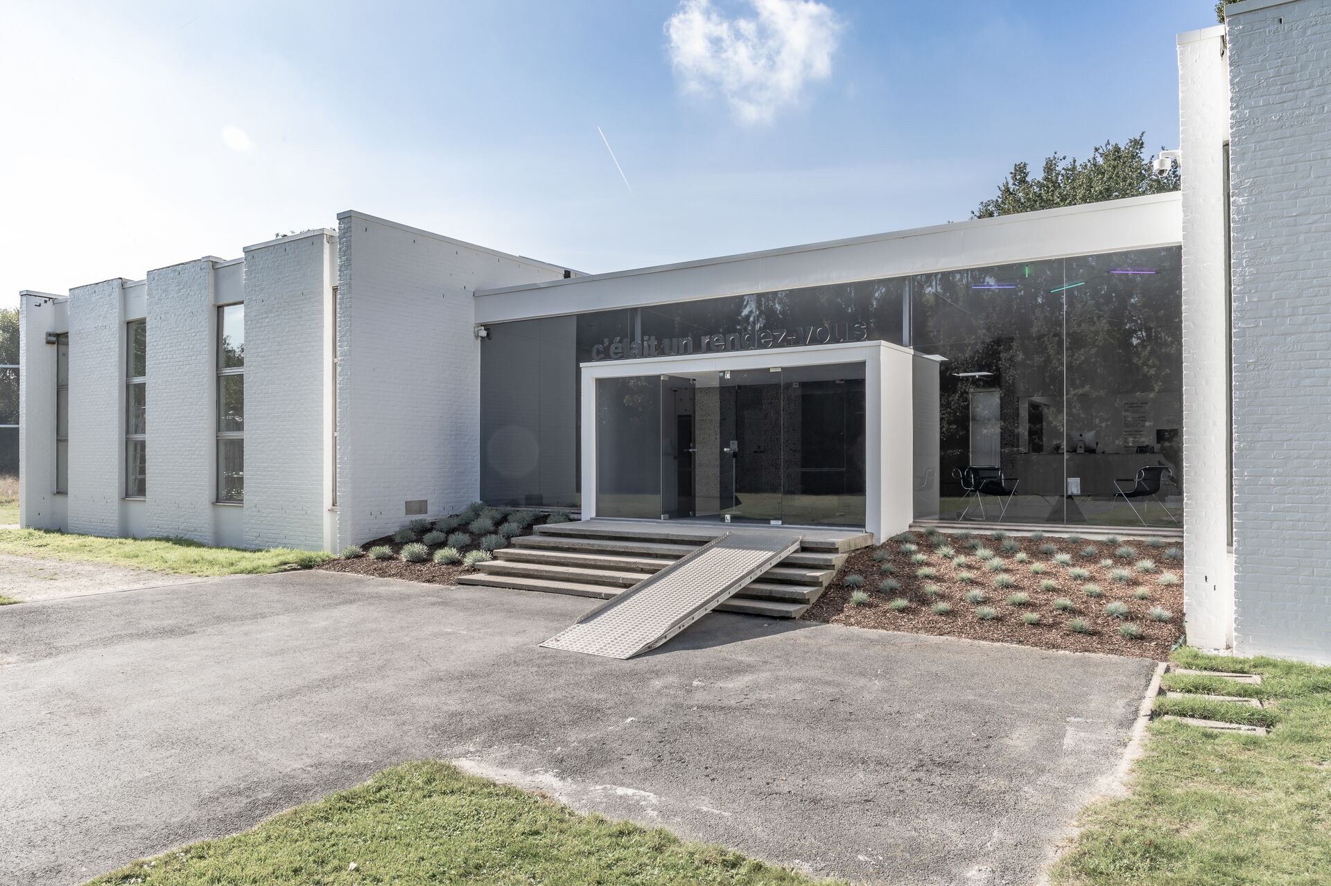 Entrada del Museum Dhondt-Dhaenens con rampa Entrada de edificio moderno blanco con fachada de vidrio, escaleras y rampa para silla de ruedas, bajo cielo azul.