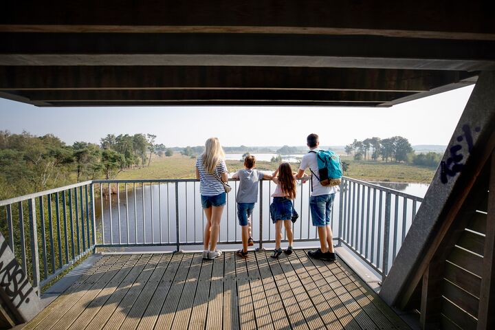 Een gezin van vier, gekleed in zomerkleding, staat op een houten platform en kijkt uit over een rustig landschap met bomen en een rivier, wat een gevoel van rust oproept.