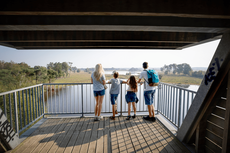 Een gezin van vier, gekleed in zomerkleding, staat op een houten platform en kijkt uit over een rustig landschap met bomen en een rivier, wat een gevoel van rust oproept.