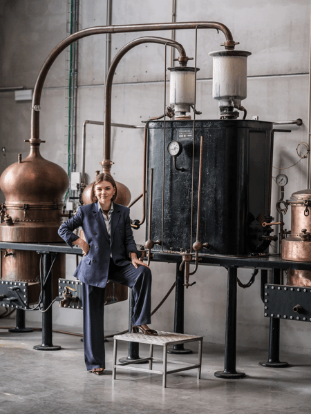 Céleste Fryn Woman in a blue suit poses by large copper stills and industrial piping in a factory hall.