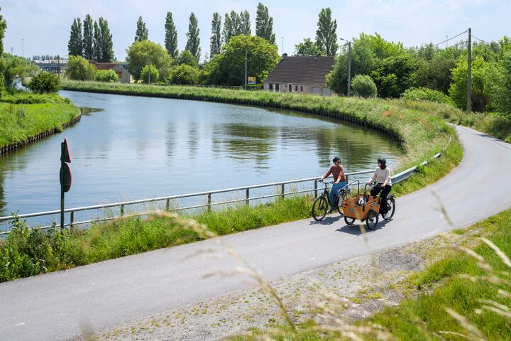 Two cyclists ride along a paved path beside a calm river, surrounded by lush greenery and trees under a clear blue sky, conveying serenity.
