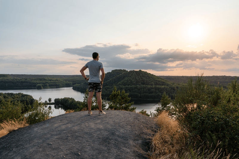 Mann steht auf einem Felsen und blickt bei Sonnenuntergang über Wald und Fluss.