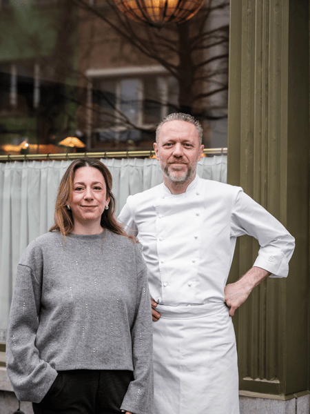 Michaël Rewers & Caroline Stasseyn Two people pose outside; a man in white chef's uniform and a woman in a grey sweater in front of a window.