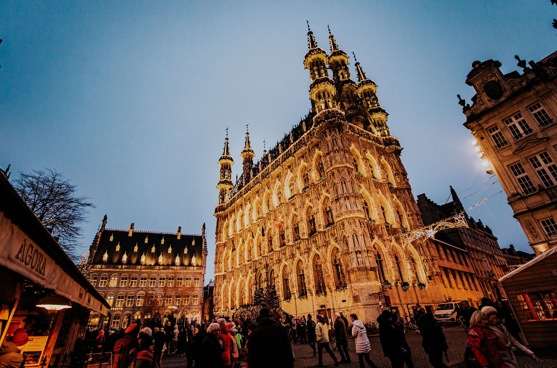 Ein großes, prachtvolles Gebäude, geschmückt mit goldenen Lichtern, dominiert die Szene vor einem Dämmerungshimmel. Menschenmengen schlendern darunter und vermitteln eine festliche, lebhafte Atmosphäre.