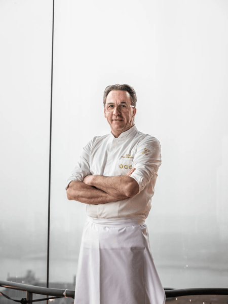 Viki Geunes Chef con uniforme blanco está de pie con los brazos cruzados frente a una gran ventana.