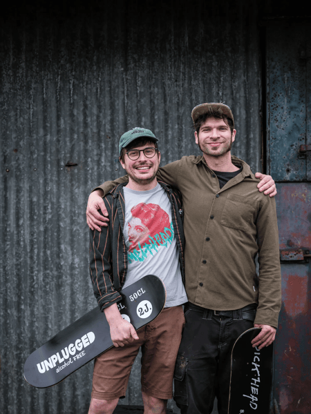 Alex Lippens & Koen Van Lancker Twee mannen met skateboard staan arm in arm voor een geribbelde metalen wand.