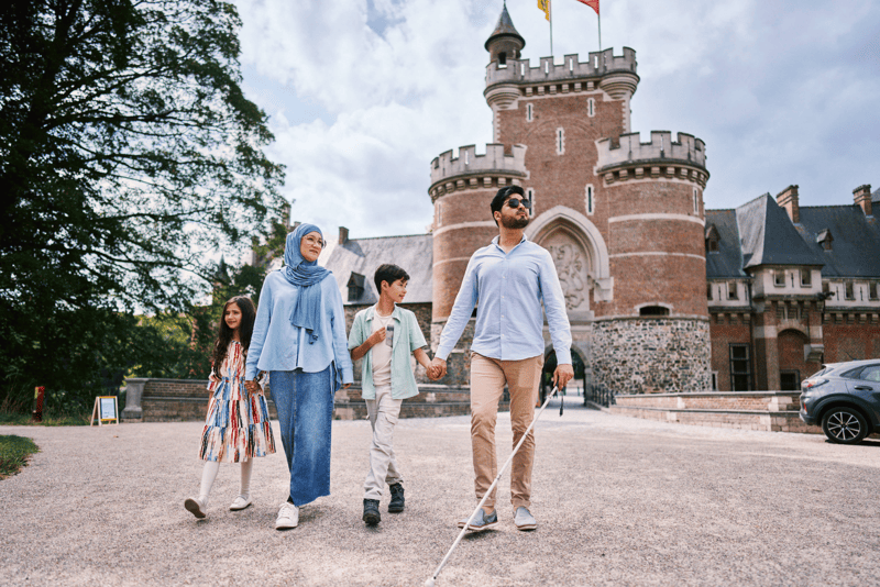 Gezin wandelt langs kasteel van Gaasbeek Gezin wandelt voor een kasteel; vader met witte stok en zonnebril houdt hand van zijn zoon vast.