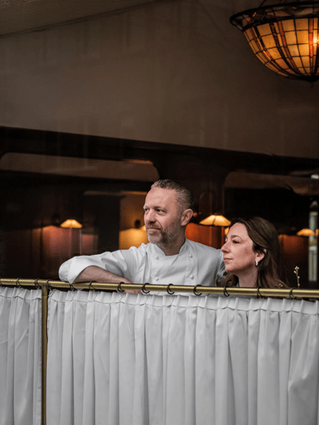 Michaël Rewers & Caroline Stasseyn Man in a white chef’s jacket and woman lean over a curtain wall in a restaurant, looking left.