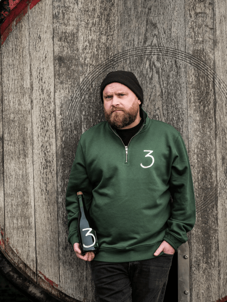 Raf Van Pottelbergh Man with beard and black beanie leans against a wooden barrel door, holds a bottle, and wears a green sweater with number 3.