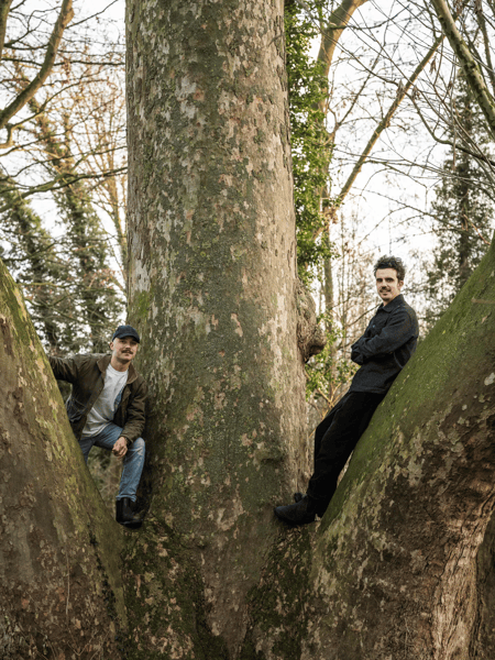 Pieter De Volder & Maxim Steijvers Two men pose between the fork of a huge tree trunk in a forest.
