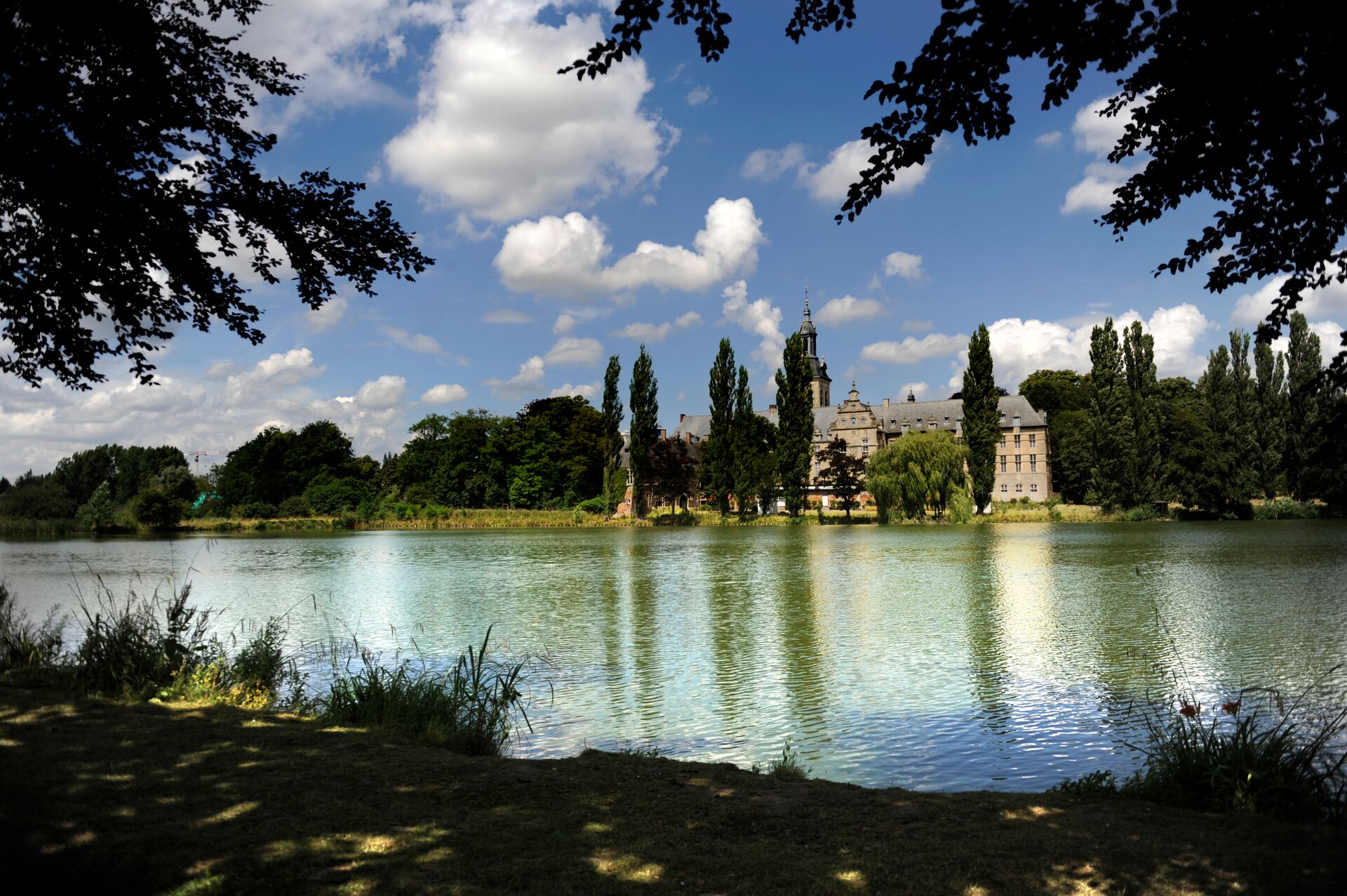 Ein ruhiges Schloss neben einem See, mit hohen Bäumen, die sich im stillen Wasser spiegeln. Blauer Himmel und flauschige Wolken schaffen eine friedliche, malerische Szenerie.