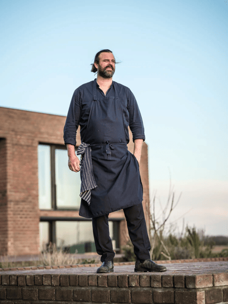 Willem Hiele Man in a dark apron stands outside on a brick edge by a modern building, with blue sky in the background.